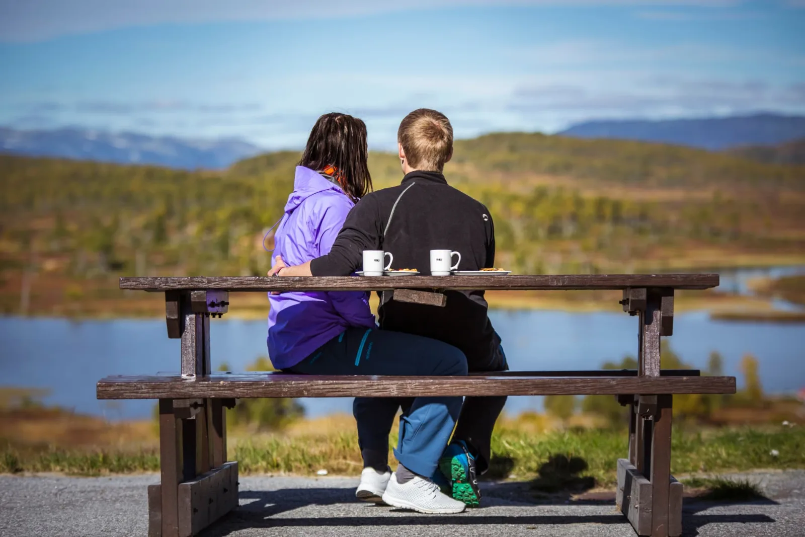 Par sitter på benk med kaffe og utsikt over fjellvann